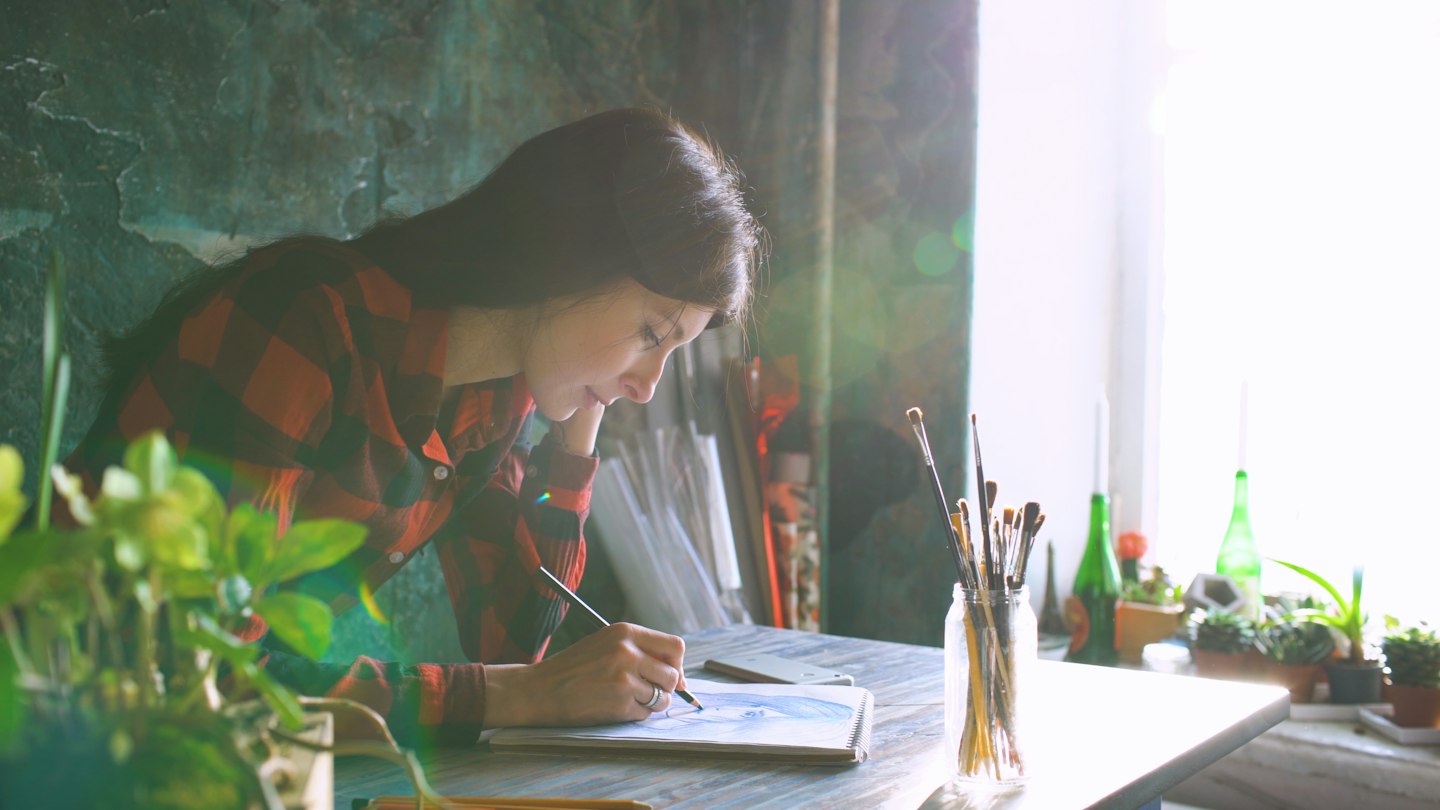 Woman painting at a desk with plants and sunlight