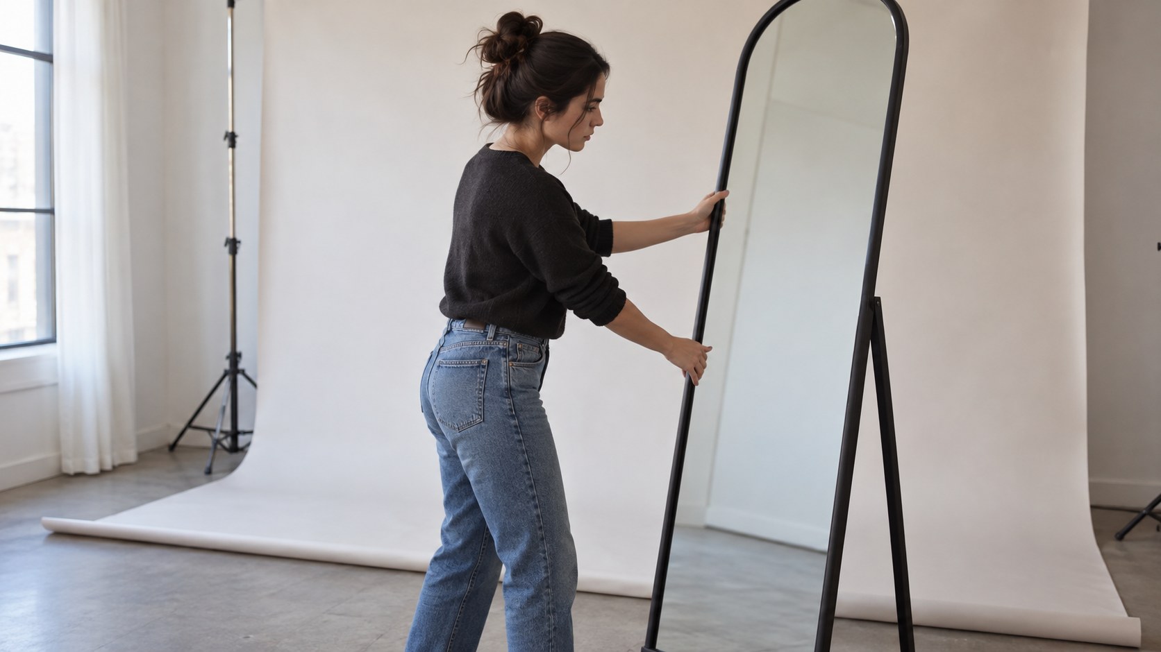 Side view of a creator moving a tall studio mirror away from a white photo backdrop while checking its reflection in natural window light.