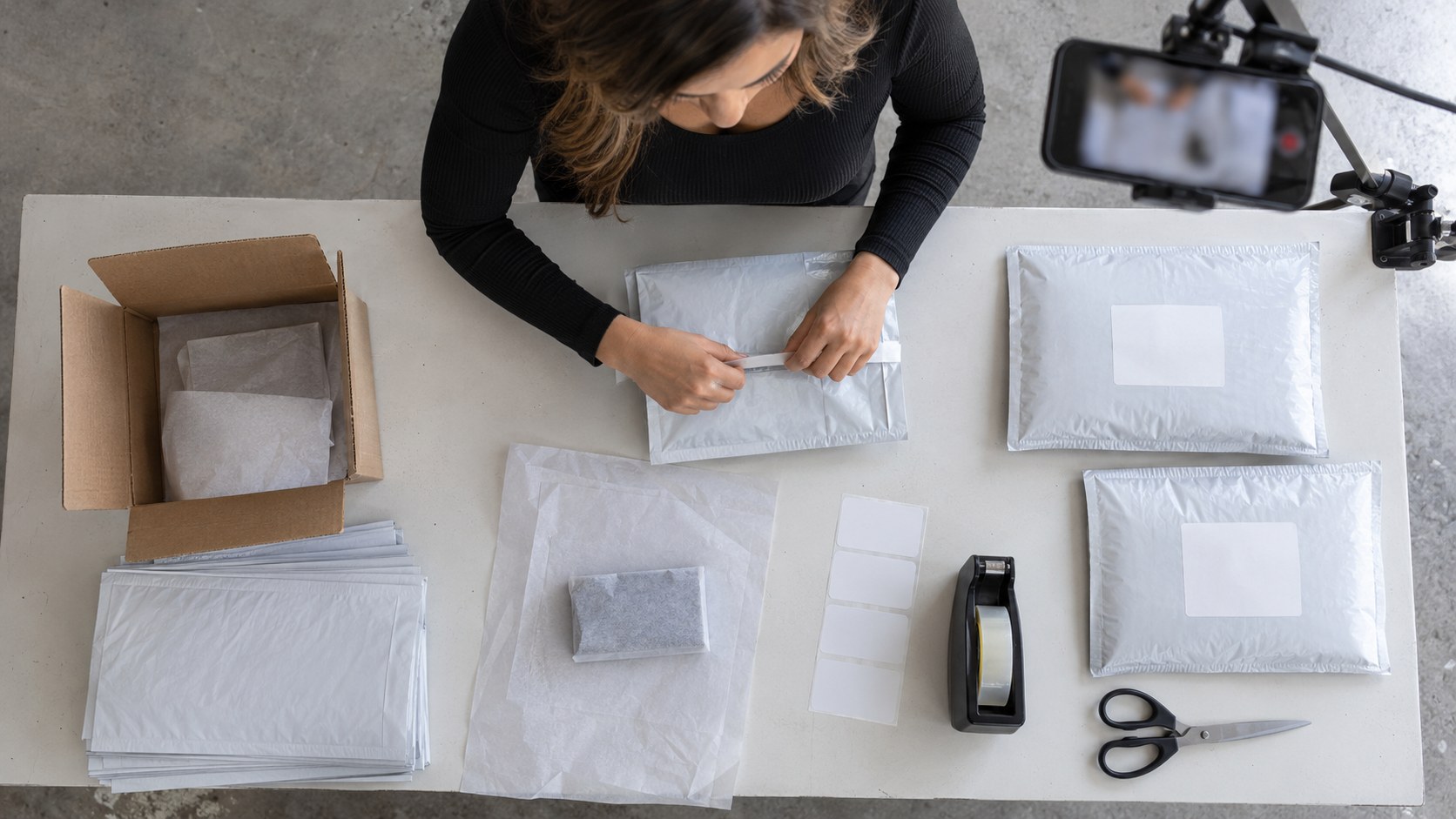 Overhead view of a female content creator sealing giveaway mailers at a studio packing table while a mounted phone records the fulfillment process.